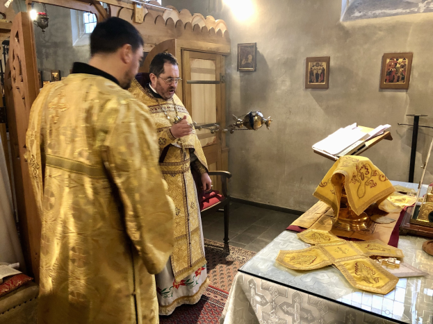 Zwei Männer in Priestergewändern stehen neben einem gedeckten Tisch mit Gegenständen in einer Kirche, mit Rahmenfotos, einem Stuhl, einer Tür und Licht im Hintergrund während einer Patriarchenweihezeremonie.