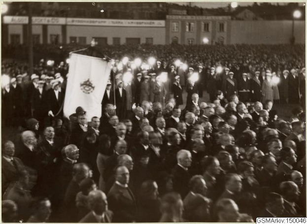 Schwarzes und weißes Bild einer großen Menschenmenge vor einem Gebäude, mit einer Person, die eine Fahne hält, in der Mitte, Pfosten und Lichter im Hintergrund und Text unten, der "Gustav Klimts Rede an der Berliner Mauer" lautet.