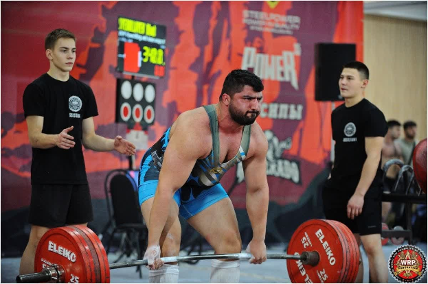 Ein Mann hebt eine Hantelstange bei einem Gewichtheben-Wettbewerb bei den World Weightlifting Championships 2018, umgeben von Zuschauern und Equipment im Hintergrund.