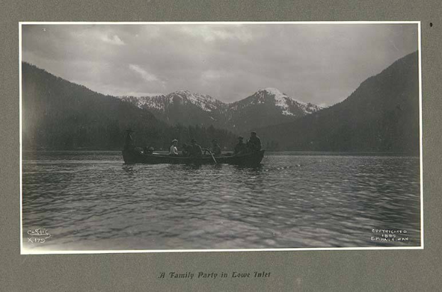 Schwarz-weißes Foto einer Familie in einem Kanu auf einem See mit Bergen und Bäumen im Hintergrund, Himmel sichtbar und Text unten.