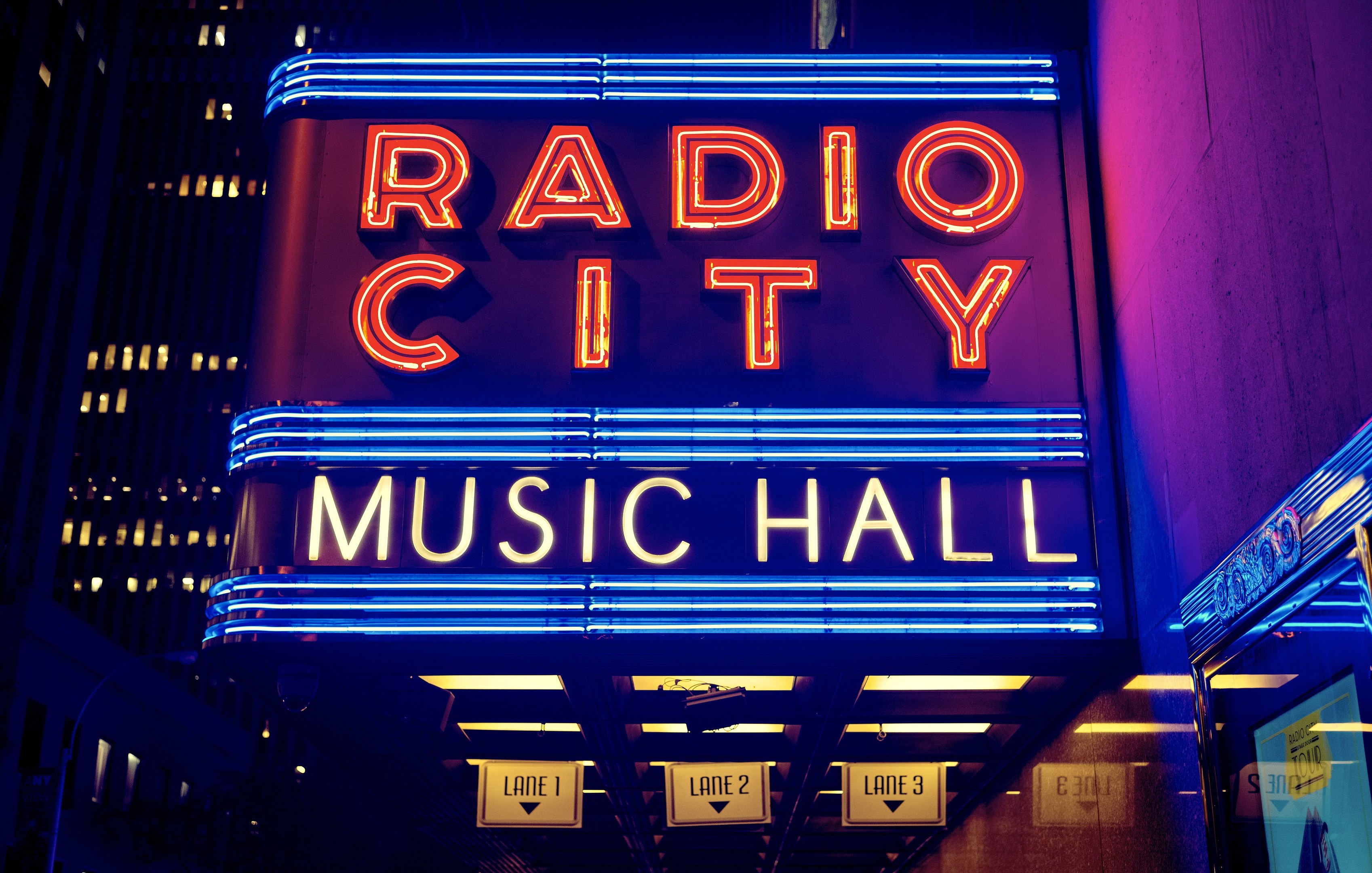 Hell erleuchtetes Radio City Music Hall in New York City mit hohen umliegenden Gebäuden und einem "Radio City"-Schild auf der rechten Seite sowie mehreren kleineren Schildern darunter.