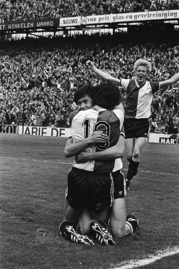 Drei Fussballspieler feiern ein Tor, indem sie sich auf dem Boden umarmen, während eine jubelnde Menge und schwenkende Fahnen im Hintergrund zu sehen sind, dargestellt in Schwarz-Weiss.