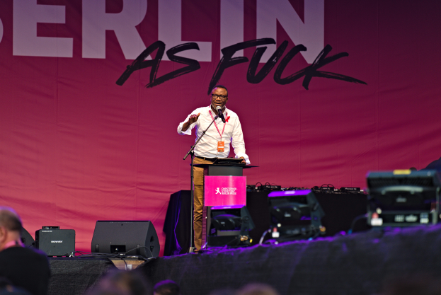 Ein Mann in einem weißen Hemd und braunen Hosen steht auf einer Bühne und hält ein Mikrofon, hinter ihm eine pinkfarbene Fahne mit der Aufschrift 'Berlin as Fuck' und Lautsprecher auf der Bühne.