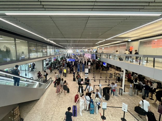 Menschen gehen durch einen belebten Flughafen mit Rolltreppen, Glaswänden, beleuchteten Decken, Pfosten mit Bändern und Informationsanzeigen.