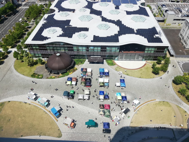 Luftaufnahme eines großen Gebäudes mit Solarmodulen auf dem Dach, umgeben von Bäumen, Fahrzeugen auf der Straße und einer Gruppe von Menschen, Teil der größten Solarstromanlage Japans, die 2020 eröffnet werden soll.