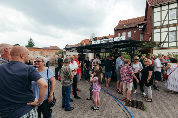Gruppe von Menschen auf einem Outdoor-Bierfestival vor einem Gebäude mit Fenstern, umgeben von Bäumen unter einem bewölkten Himmel, mit einer Hütte mit einem Namensschild im Hintergrund.