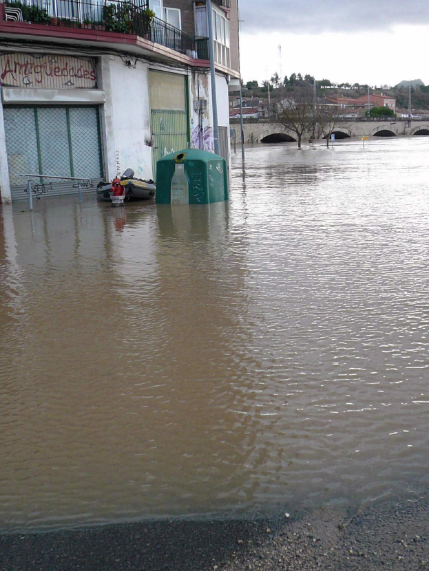 Überschwemmte Straße mit einem von Wasser umgebenen Gebäude, ein Nachbargebäude mit Fenstern, Geländern, Pflanzen und einer Namensplakette sowie ein Hintergrund mit Bäumen, einer Brücke, Pfählen und anderen Gebäuden sowie einem bewölkten Himmel.