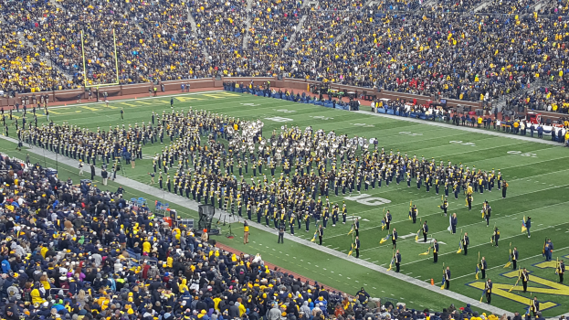Eine große Menschenmenge in einem Stadion bei einem Fußballspiel, wobei einige stehen und Fahnen halten, andere sitzen und verschiedene Gegenstände wie Pfosten und Geländer im Hintergrund zu sehen sind.