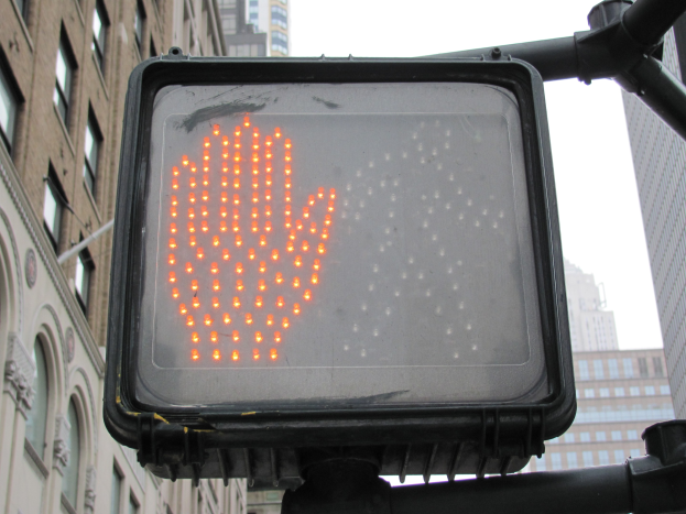 Eine Ampel mit rotem Handzeichen befindet sich in der Mitte einer Stadt, flankiert von Gebäuden auf beiden Seiten unter einem klaren Himmel.