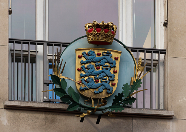 Wappen der britischen Königsfamilie, das auf der Außenseite eines Gebäudes dargestellt wird, begleitet von einem Logo mit Krone und Blättern sowie Fenstern und Gittern im Hintergrund.