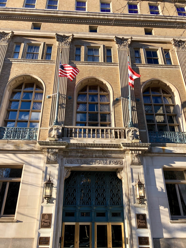Außenansicht des Plaza Hotels in New York City mit Glasfenstern, -türen, -säulen, -geländern, -leuchten, Texttafeln und Flaggen vor einem Himmel-Hintergrund.