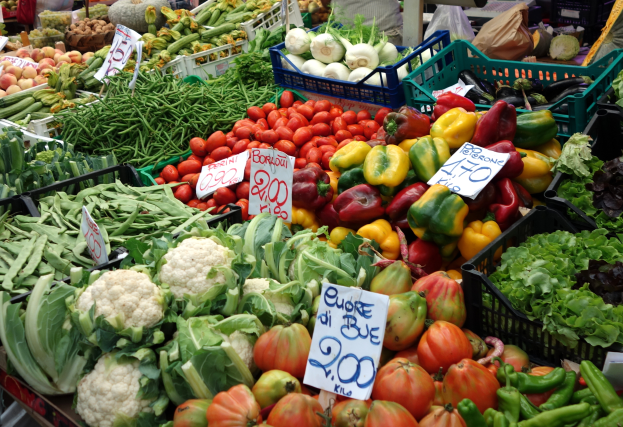Ein belebter Bauernmarkt mit verschiedenen Gemüsesorten wie Tomaten, Paprika, Blumenkohl, grüne Bohnen in Körben, Preisetiketten sichtbar und einige Menschen schlendern.