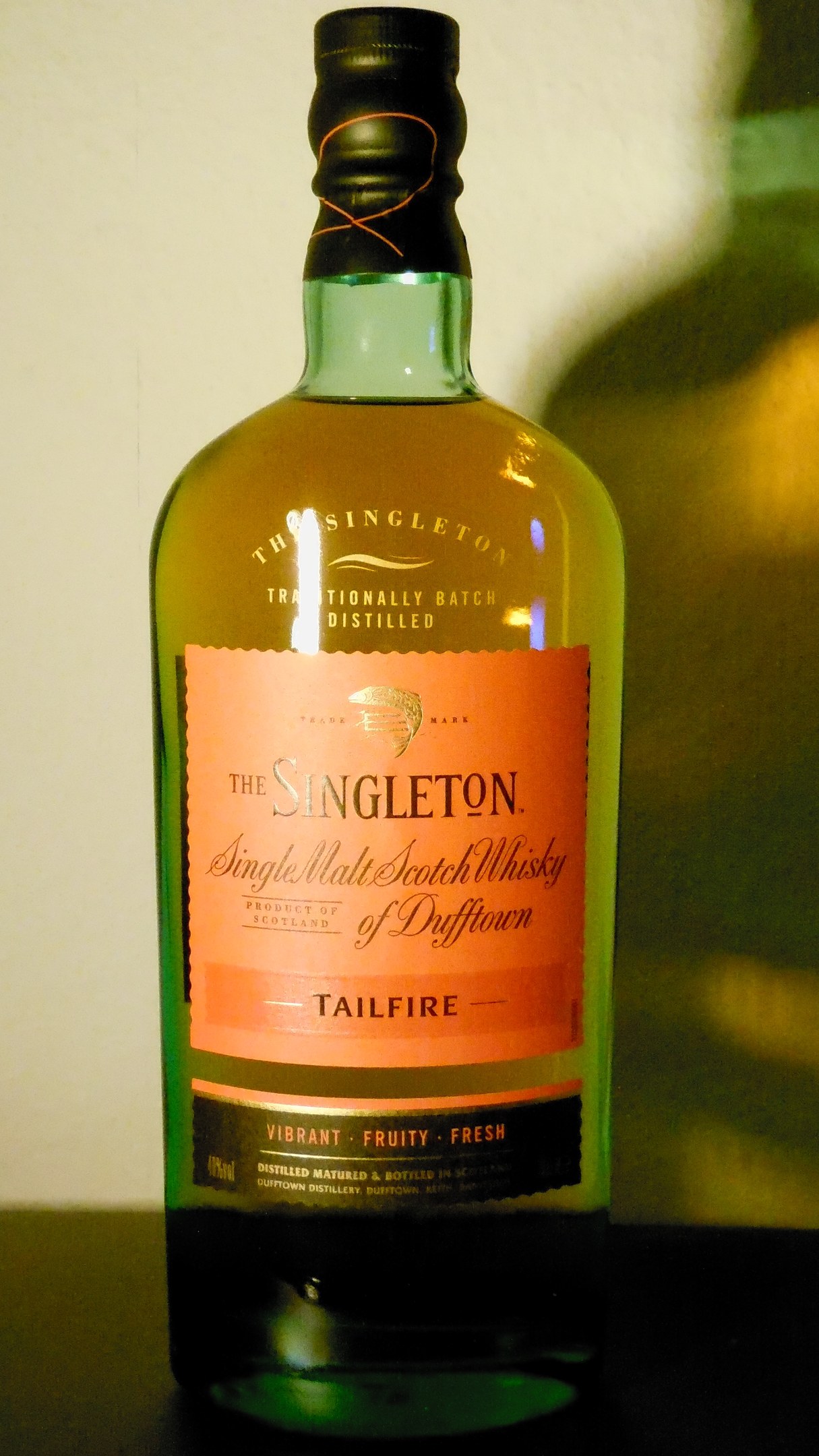 Dunkle Bernsteinflasche von Singleton of Dufftown Tailfire Single Malt Scotch Whisky mit einem schwarzen Etikett, das ein Löwen- und ein Burgwappen zeigt, auf einem Tisch.