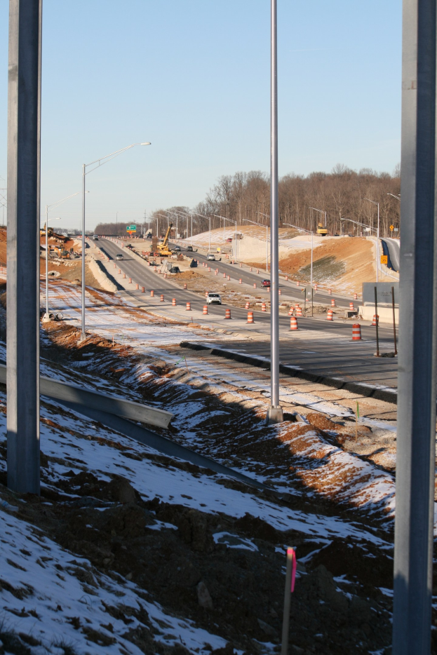 Baustelle mit schneebedecktem Boden, Pfählen, Absperrkegeln, Straßenlaternen, Baumaschinen, Bäumen und einem klaren Himmel im Hintergrund.
