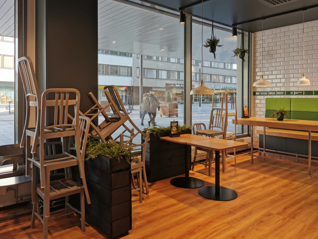 Ein Berliner Restaurant mit Holztischen und -stühlen, sichtbar durch ein großes Fenster umgeben von Pflanzen und Lichtern, mit Passanten und Gebäuden draußen.