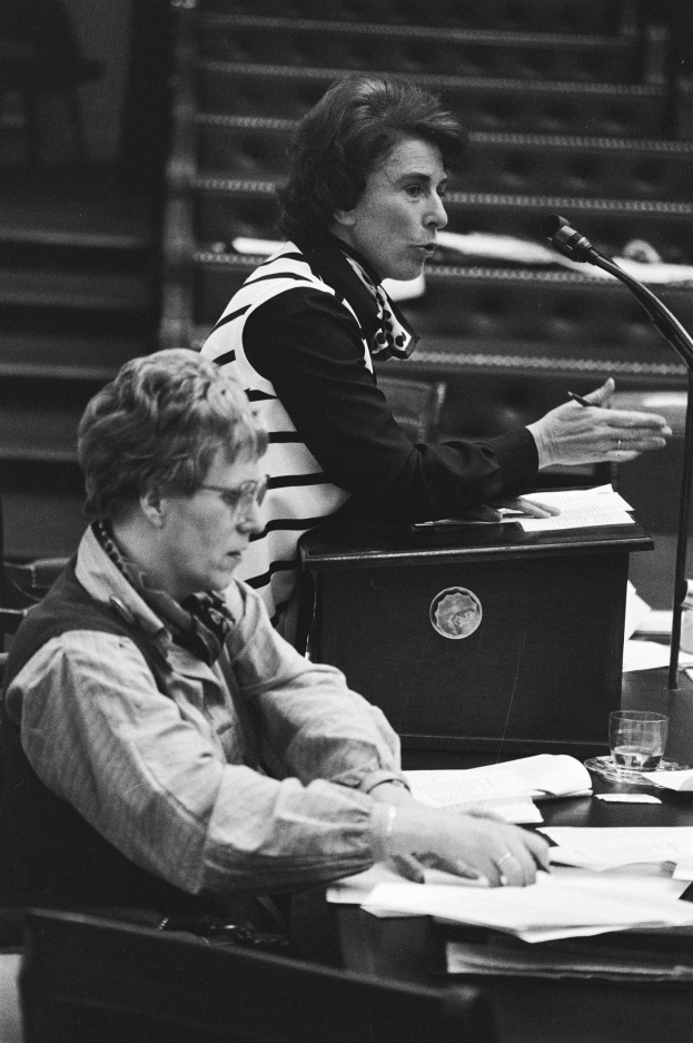 Schwarz-weißes Foto einer Frau, die an einem Tisch im Europäischen Parlament in Straßburg im Mai 1981 sitzt und einen Stift hält, mit Papieren, einem Glas Wasser und anderen Gegenständen auf dem Tisch, mit Stufen im Hintergrund.