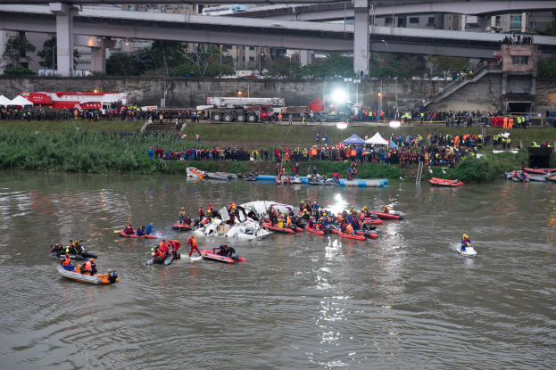 Eine Gruppe von Menschen in kleinen Booten, die Lebenswesten tragen, treiben auf dem Wasser, umgeben von Pflanzen, Gras, Zelten, Fahrzeugen, Laternenmasten, Bäumen, Gebäuden und einer Brücke, was auf einen Rettungseinsatz hinweist.