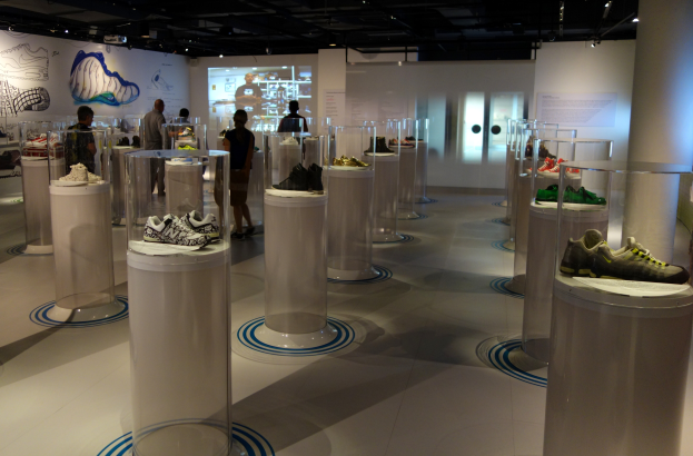 Ein Raum voller verschiedener Schuharten, die in Glasboxen ausgestellt sind, mit Menschen auf dem Boden, einem Bildschirm an der Wand und einer beleuchteten Decke, wahrscheinlich im Nike-Museum in New York City.