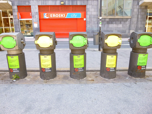 Eine Reihe von Parkautomaten entlang einer Straße mit einem Gebäude mit Glasfenstern und einer Rolllade mit Text im Hintergrund und einem nahen Müllcontainer.
