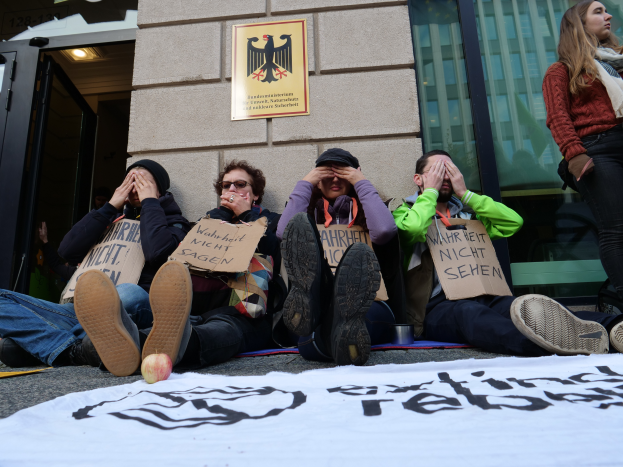 Eine Gruppe von Menschen sitzt vor einem Gebäude auf dem Boden und hält Schilder hoch, mit einem Banner und einem Apfel auf der Straße und einer Frau, die rechts steht und besorgt aussieht.