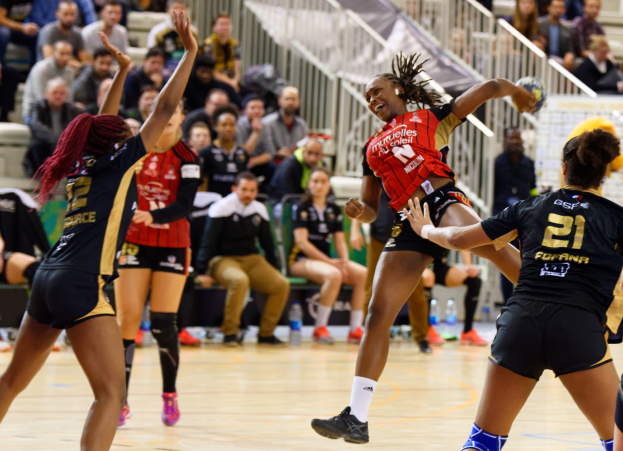 Eine Gruppe von Frauen, die Volleyball auf einem Feld mit einem Ball in der Mitte spielt, während Zuschauer im Hintergrund auf Stühlen sitzen, Geländern und Flaschen auf dem Boden.
