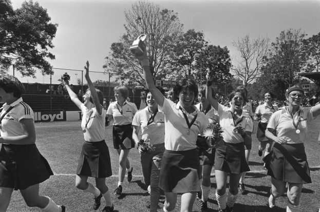Eine Gruppe von Frauen rennt auf einem Rasenfußballfeld, jede hält einen Pokal in der Hand, mit Bäumen, Maschendrahtzaun und Texttafeln im Hintergrund, dargestellt in Schwarz-Weiß.