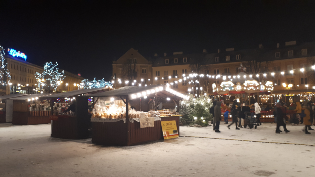 Ein verschneiter nächtlicher Weihnachtsmarkt mit Menschen, Buden mit Lichtern, Pflanzen, Bäumen, Gebäuden und Schildern unter einem bewölkten Himmel.