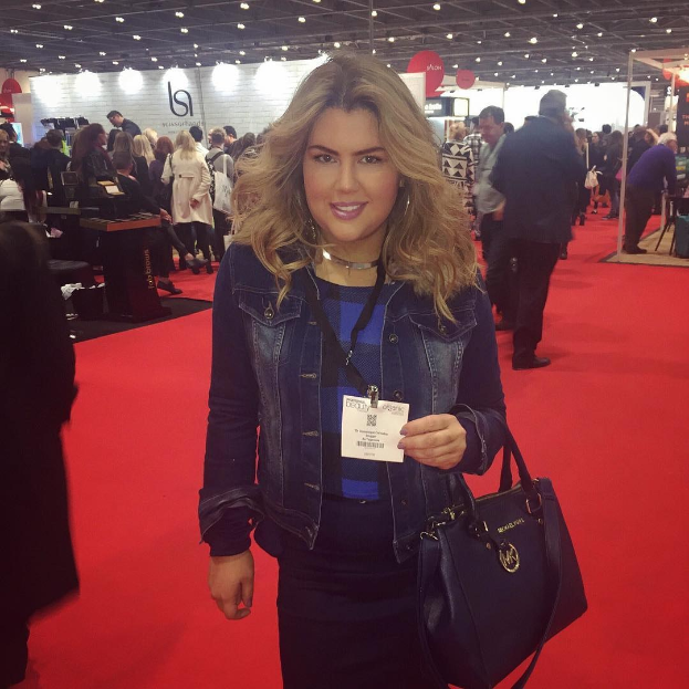 Eine Frau in einem roten Kleid steht auf einem roten Teppich bei einer Modenschau, lächelt und hält eine Handtasche, während Fotografen und Scheinwerfer im Hintergrund zu sehen sind.