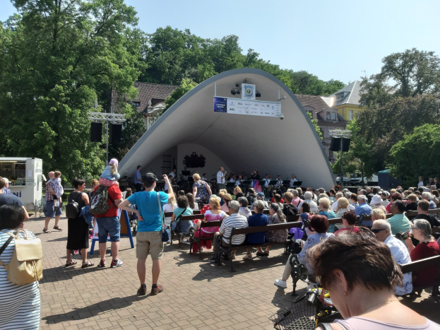 Menschen auf Bänken vor einer Bühne mit Stehenden, einer Kamera, einem Stativ, einem Banner, Pfosten, einem Metallrahmen, Bäumen, Gebäuden mit Fenstern und einem bewölkten Himmel bei einem Sommerkonzert im Park.