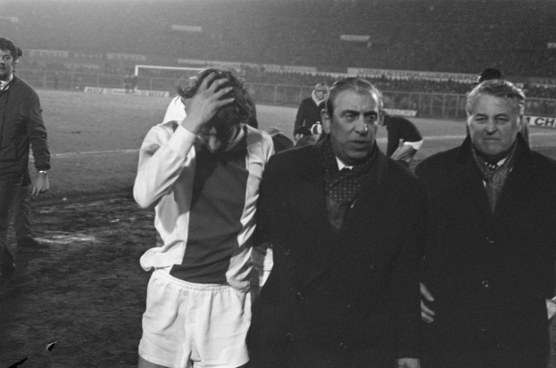 Eine Gruppe von Männern in Not steht zusammen auf einem Fußballfeld, mit einem Stadion im Hintergrund, auf einem Schwarz-Weiß-Bild.
