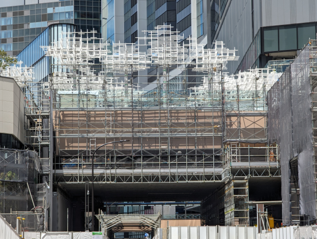 Großes Gebäude mit Gerüst, umgeben von Bäumen links und einer Straße unten in einer städtischen Umgebung.