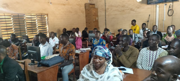 Eine Gruppe von Menschen sitzt an Tischen in einem Klassenzimmer mit Monitoren, Taschen, Büchern und Papieren auf den Tischen, ein Fenster auf der linken Seite und Kartons und einen Fernseher an der Wand im Hintergrund, an einer Schulung zur Technologie-Nutzung teil.