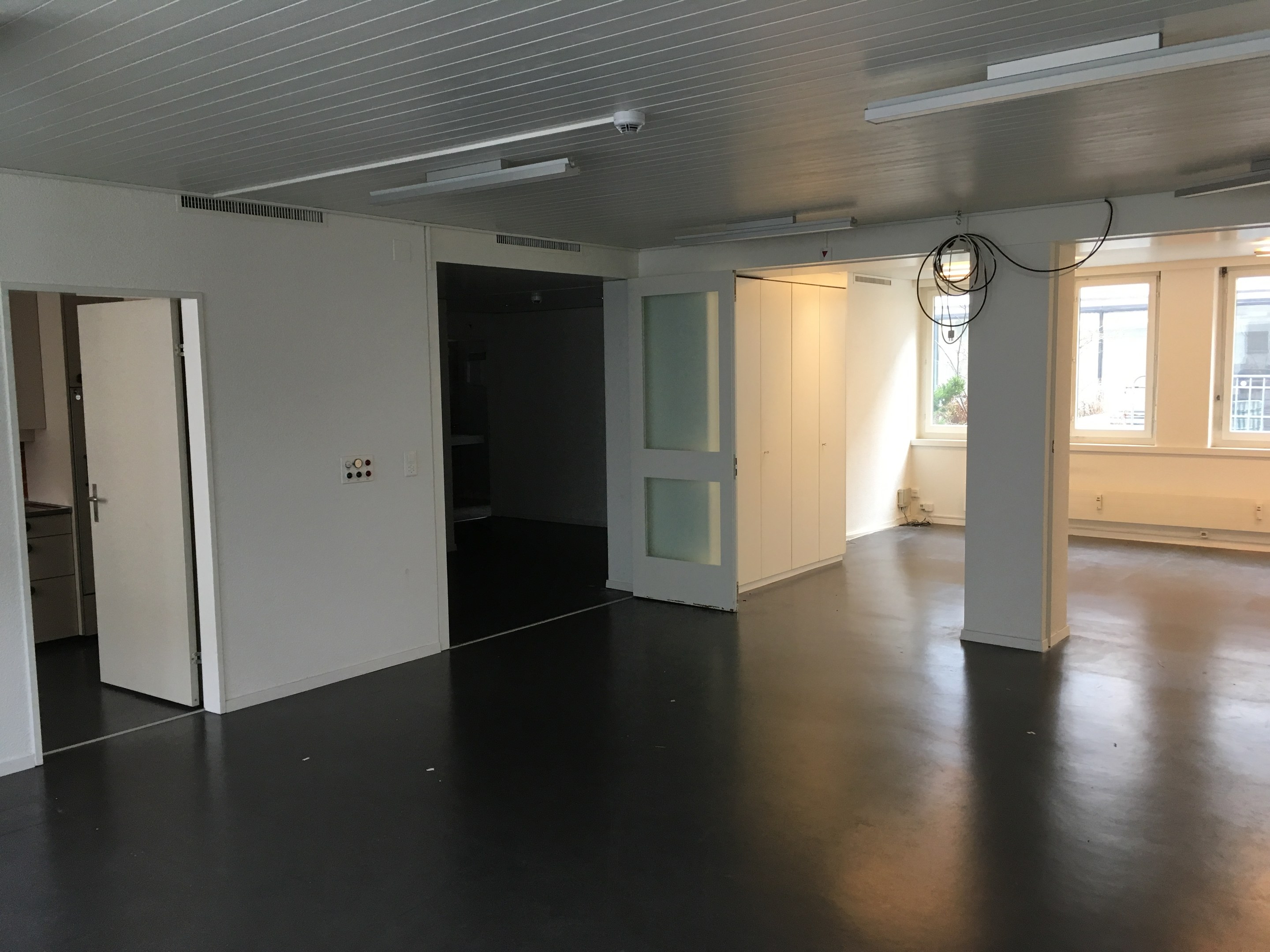 Leerer Büroraum mit schwarzen Böden, weißen Wänden, Deckenleuchten, sichtbaren Kabeln, Türen, Schränken, Fenstern und einer Außenansicht von Gebäuden und Bäumen in Amsterdam.