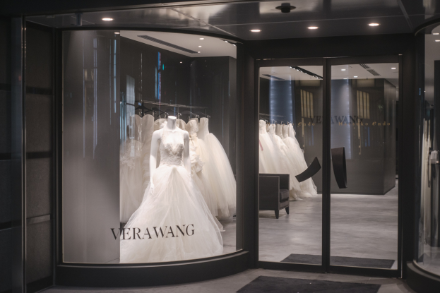 Innenansicht eines Brautmodengeschäfts in Singapur mit Hochzeitskleidern auf Schaufensterpuppen und Bügeln, einer Glaswand, einem Teppich, einem Stuhl und Deckenleuchten.
