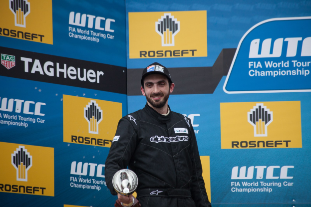 Ein Mann in einem schwarzen Kleid und einer Mütze steht auf einem Podium, lächelt und hält eine Trophäe in der Hand, mit einer blauen Tafel im Hintergrund, die "FIA World Touring Car Championship" trägt.