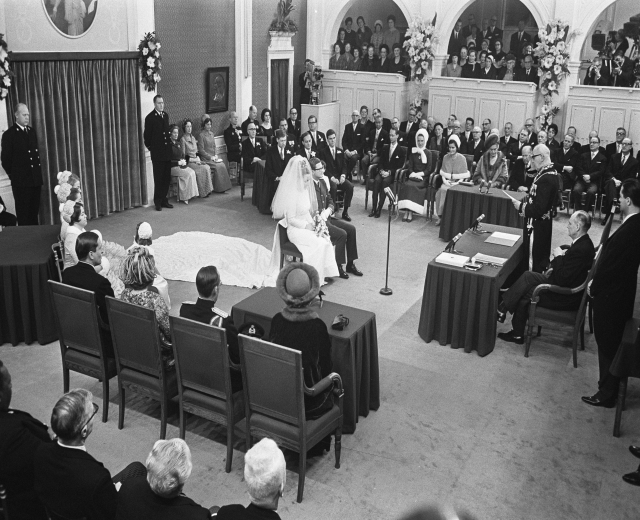 Ein Schwarz-Weiß-Foto einer Hochzeitszeremonie in einem großen Raum mit sitzenden und stehenden Gästen, Tischen mit Mikrofonen und Papieren, einer Wand mit Rahmenfotos, Blumenbouquets, einem Vorhang und einem Podium mit Mikrofonständer.