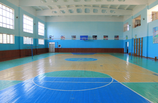 Eine große Turnhalle mit einem blauen und weißen Boden, die einen Basketballplatz, ein Netz, Stangen, eine Tür, Fenster, Plakate an der Wand, ein Geländer und ein Dach zeigt.