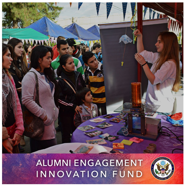 Gruppe von Menschen um einen Tisch versammelt mit einer "Alumni Engagement Innovation Fund"-Aufschrift, umgeben von Gegenständen, mit Zelten, Bäumen und einem klaren Himmel im Hintergrund, und Text und ein Logo unten.