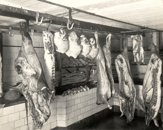 Ein Schwarz-Weiß-Foto eines Metzgergeschäfts mit verschiedenen Fleischstücken, die von der Decke hängen, einer Wand im Hintergrund und einer Decke oben.