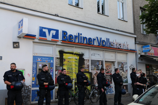 Polizisten vor einer Berliner Volksbank stehend, einige mit Helmen, daneben ein Auto und ein Baum, Gebäude und Texttafeln im Hintergrund.