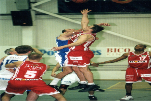 Eine Gruppe von Männern, die Basketball auf einem Court mit einer Wandlautsprecher und Text im Hintergrund spielen, mit einem Basketball in der Luft darüber.