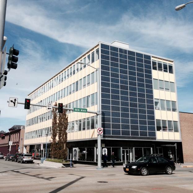 Großes Gebäude mit Solarpaneelen an der Seite, umgeben von Straßeninfrastruktur einschließlich Pfählen, Laternen, Signalen und Schildern, mit Kraftfahrzeugen, Fußgängern, Bäumen und einem bewölkten Himmel im Hintergrund.