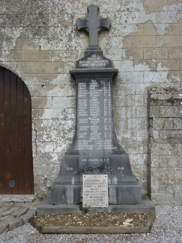 Ein steinernes Denkmal mit einem Kreuz, das eine Tür auf der linken Seite hat, vor einer Steinwand mit einer Basis aus verstreuten Steinen steht.