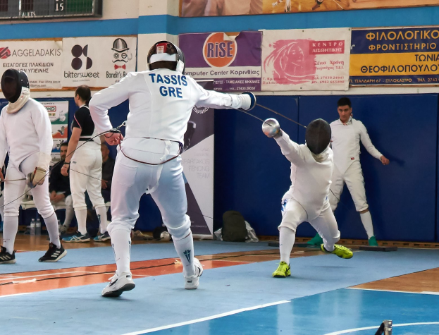 Gruppe von Menschen, die in einer Turnhalle mit Helmen und Schwertern ficht, mit einer Anzeigetafel darüber und Zuschauern auf Stühlen im Hintergrund.