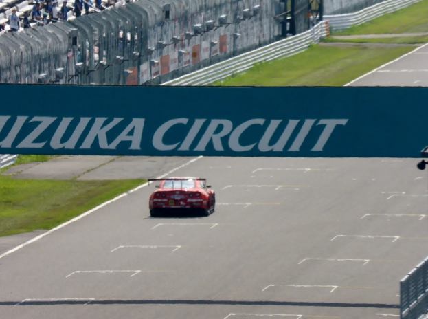 Roter Sportwagen auf einer Rennstrecke mit "Suzuka Circuit"-Schilder im Hintergrund, umgeben von Gras, Absperrungen und Zuschauern auf den Tribünen.