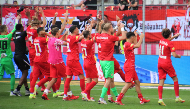 Eine Gruppe von Fußballspielern, die ein Tor feiern, auf einem Feld mit einigen, die Flaschen halten und andere klatschen, vor einem Hintergrund aus einem Zaun, Bannern, Metallstangen, Stühlen und einer Decke mit Deckenleuchten.