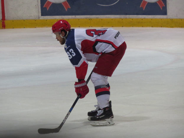 Ein Hockey-Spieler in voller Ausrüstung, einschließlich Helm und Handschuhen, hält einen Stock, während er auf dem Eis fährt, mit einem Banner mit Text und Logos im Hintergrund.