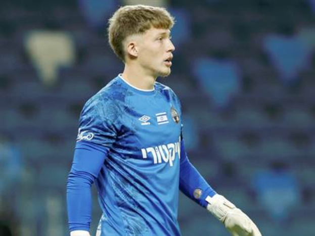 Ein junger Mann in einem blauen Torwarttrikot und weißen Handschuhen steht auf einem Fußballfeld während eines Spiels, mit einem unscharfen Hintergrund.