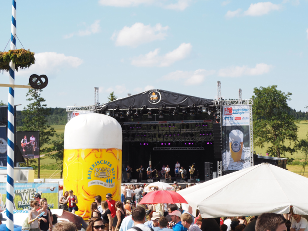Eine große Menge beim Oktoberfest in München, Deutschland, steht vor einer Bühne mit Musikern, viele tragen Mützen, Brillen und halten Schirme, mit einem dekorierten Pfahl und einem Zelt im Vordergrund.