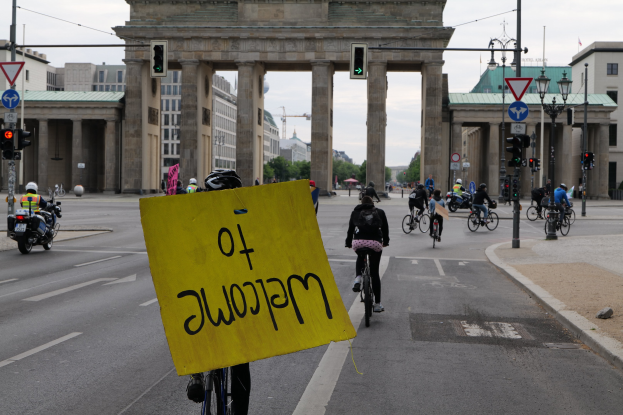 Eine Gruppe von Radfahrern fährt am Brandenburger Tor in Berlin vorbei, einer hält ein gelbes Schild, mit Laternenmasten, Ampeln, Gebäuden, Bäumen und einem klaren Himmel im Hintergrund.